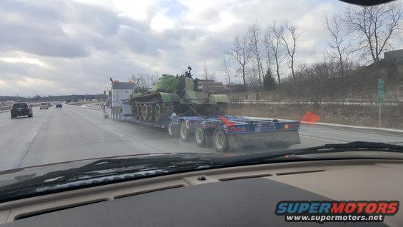Green tank on a blue flatbed trailer being transported on a highway