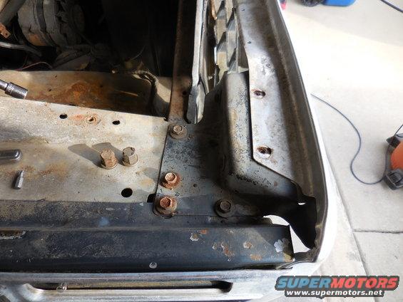 Close-up of a rusty metal bracket on a vehicle's fender mounting area, showing bolts and some engine components in the background
