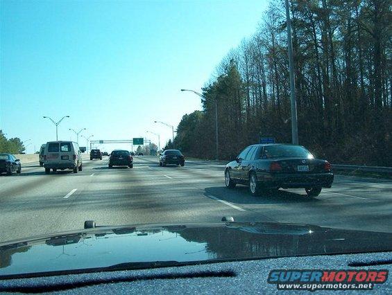 Black sedan driving on highway, rear view. License plate is partially visible