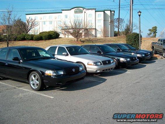 Five Ford Crown Victoria police interceptors parked in a lot, one black, four dark colors. 2001 Ford Crown Victoria