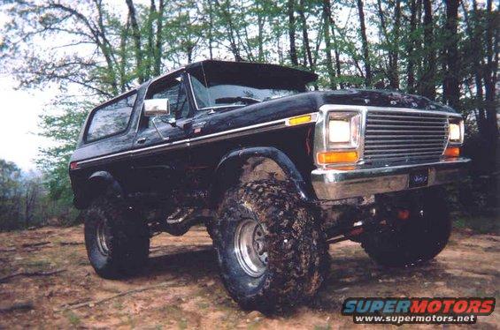 Black 1978 Ford Bronco with large off-road tires parked on dirt with trees in background