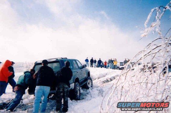 People pushing a dark SUV stuck in deep snow, with more people and vehicles in the background