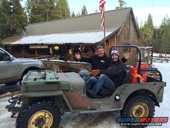 Old Jeep Willys MB with people in it, parked outside a building with snow on the roof. Another vehicle is to the left
