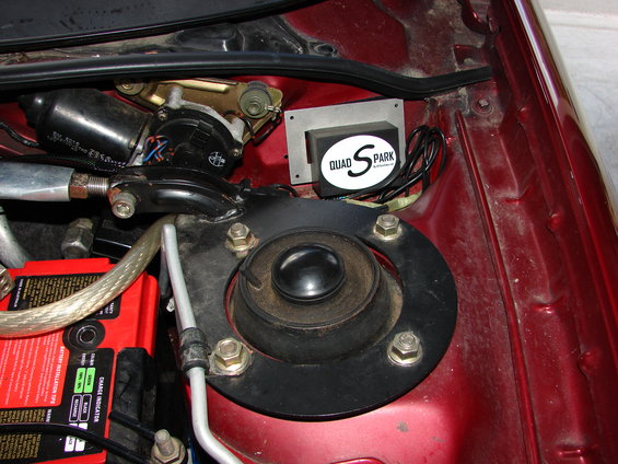 Close-up of car's engine bay showing a Quad Spark ignition module, battery, and windshield wiper motor