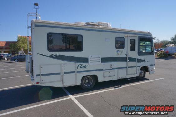 RV parked in a lot, side view. It has a ladder on the back and a 'Flair' decal