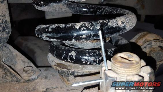 Close-up of a dirty black coil spring with a metal pin inserted, resting on a metal mount with a large nut