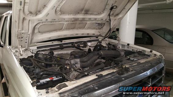White SUV engine bay with hood open. Visible engine components and battery. Watermark "Supermotors.net" at bottom right