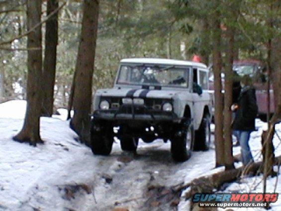 Light-colored SUV with black racing stripes on the hood, driving in snow. Front grille and headlights are visible