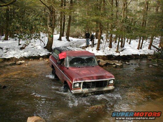Red Ford Bronco driving through a shallow stream in a wooded area with snow on the ground