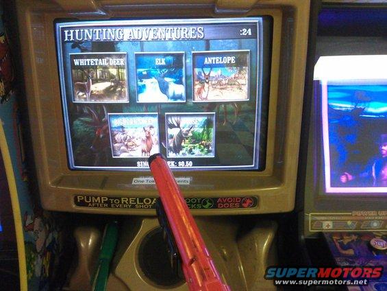 Arcade game screen showing 'HUNTING ADVENTURES' with options for Whitetail Deer, Elk, and Antelope. A red toy gun is in the foreground