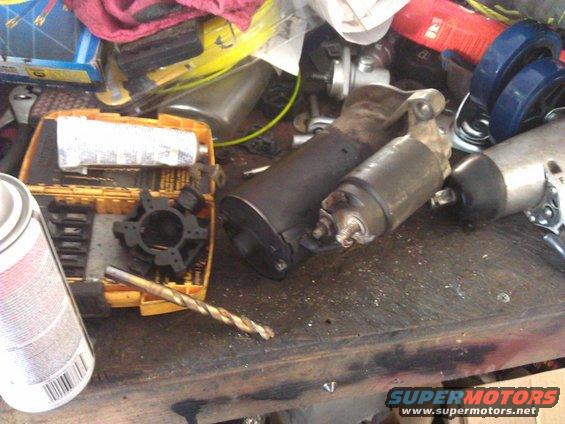 Two automotive starters and a drill bit on a workbench with tools