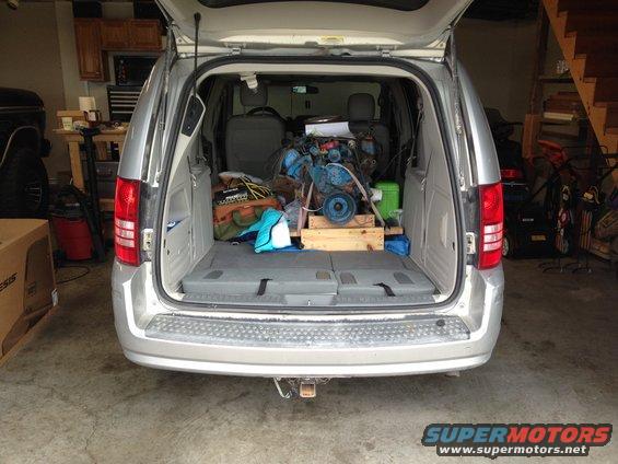 Silver minivan rear view with open trunk containing a blue engine on a wooden stand