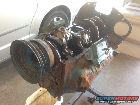 Rusty engine block with crankshaft and pulleys, partially disassembled, on a stand. A white car is visible in the background