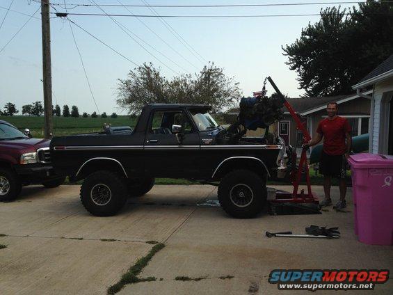 Black 1978 Ford Bronco with hood open, engine being lifted out by an engine hoist. Another vehicle is parked to the left
