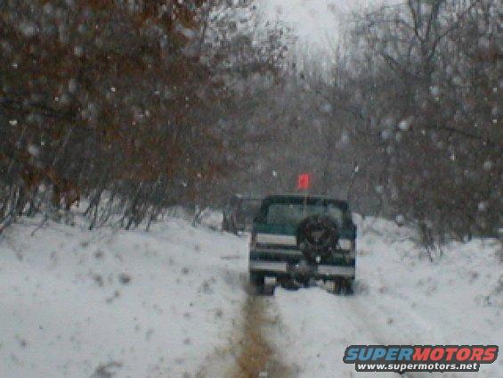 Two SUVs drive on a snow-covered road with trees on either side. One SUV has a spare tire mounted on the back and a red flag on top