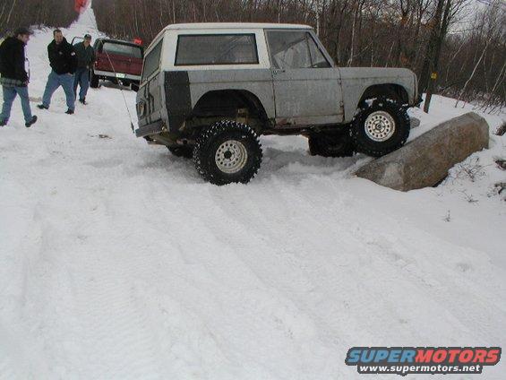 Off-road vehicle with large tires navigating a snowy incline with a large rock. Another vehicle and people are visible in the background