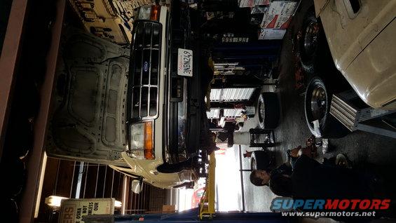 Ford F-Series truck on lift, hood open, mechanic working underneath. License plate 3NBN249