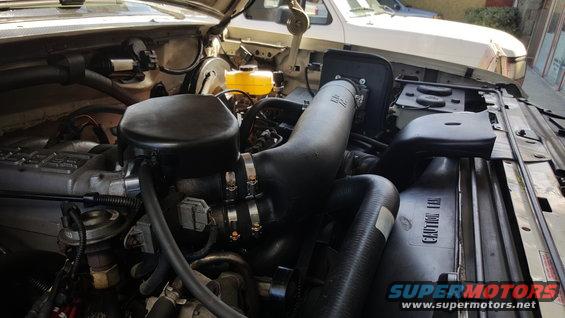 Engine bay of a 1995 Ford Bronco with aftermarket air intake. The engine is visible, along with hoses and other components. A second white vehicle is partially visible in the background