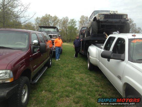 Several trucks are parked on grass, one is a flatbed hauling a Jeep. Two men are standing around
