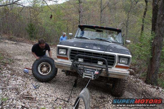 Man pushing a spare tire next to a black Ford Bronco with a winch on the front. Another person stands behind the vehicle