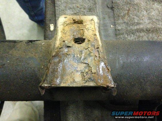 Close-up of a heavily rusted metal bracket attached to a dark, cylindrical axle. The bracket has a trapezoidal shape with a hole in the top