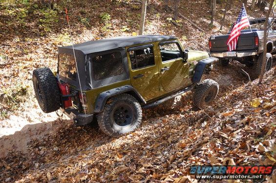Olive green Jeep Wrangler and a pickup truck with an American flag drive on a leaf-covered dirt trail