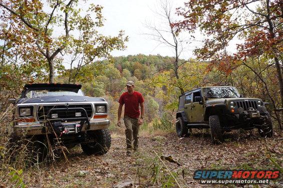 Ford Bronco and Jeep Wrangler parked on a dirt path in autumn woods. Man walks between them