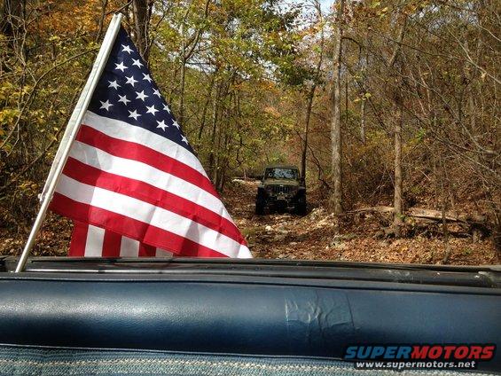 American flag waving from a vehicle, with another Jeep on a wooded dirt road ahead