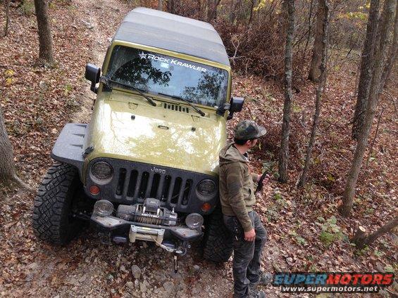 Yellow Jeep Wrangler parked on a wooded trail with a person leaning against it. Decal on windshield reads 'Rock Crawler'