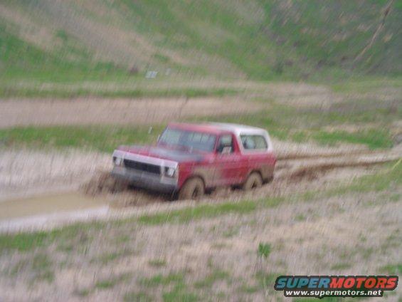 Red 1978 Ford Bronco stuck in mud, front-left wheel submerged. Off-road scene with muddy track