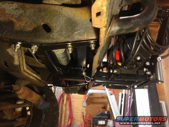 Underside view of a vehicle frame with a new winch mount being installed. Shows bolts, cables, and part of a tire