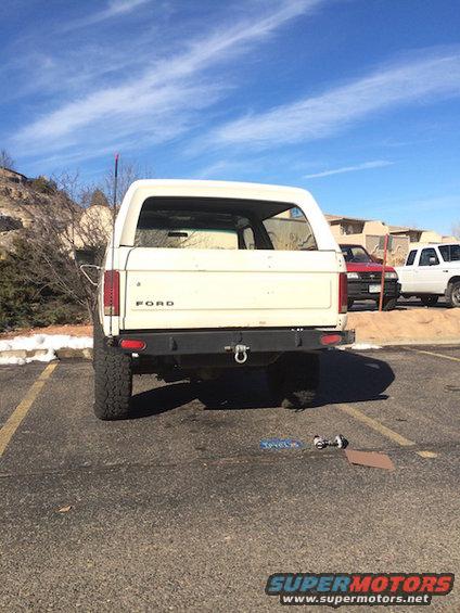 White Ford Bronco parked in a lot, rear view. Another red car and white truck are visible