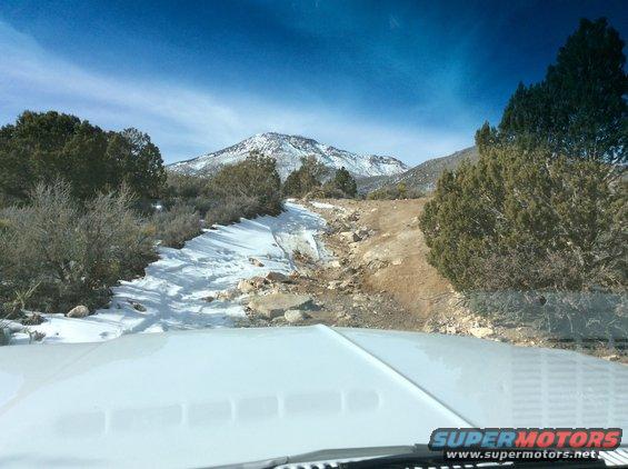 Driving on a snowy, rocky trail towards mountains under a blue sky. Part of a white vehicle hood is visible