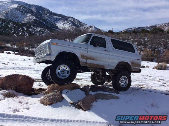 Lifted white 1978 Ford Bronco with chrome wheels on rocks in a snowy landscape