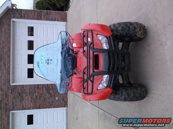 Red ATV with a clear windshield parked in front of a brick garage with white doors. The windshield has graphics and the word HONDA