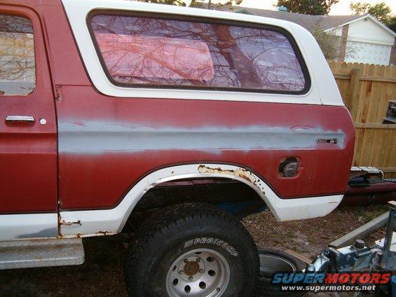 Side view of a red and white Ford Bronco with primer on the rear quarter panel. Gas cap door is open