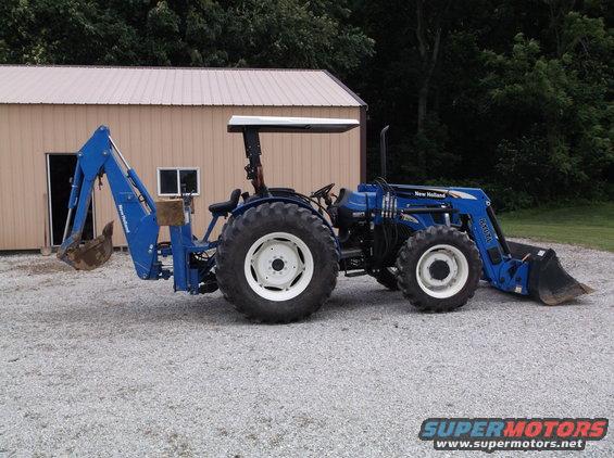 Blue New Holland tractor with front loader and backhoe attachment parked on gravel in front of a shed