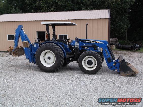 Blue New Holland tractor with front loader and backhoe attachment parked in front of a building