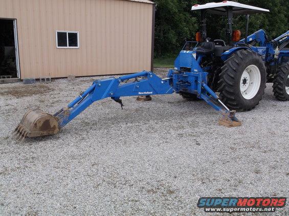 Blue tractor with a New Holland backhoe attachment, viewed from the side with the bucket on the ground