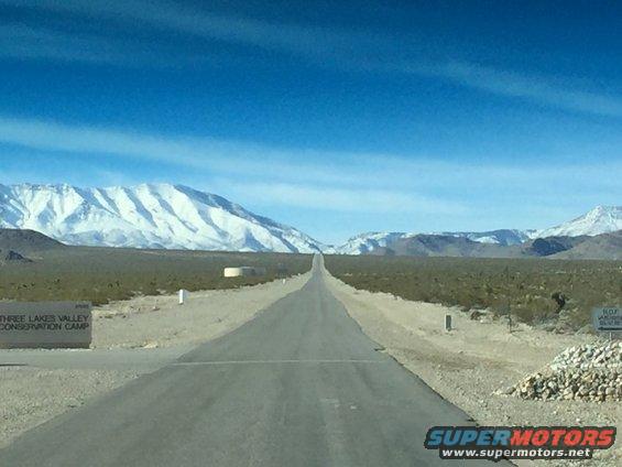 Desert road leading to snow-capped mountains, sign for Three Lakes Valley Conservation Camp