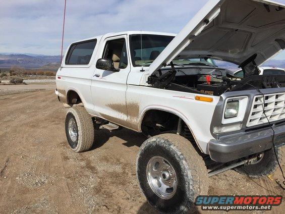 White 1978 Ford Bronco with hood open, covered in mud, parked on a dirt road