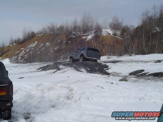 Two SUVs driving on a snowy, hilly terrain with bare trees in the background