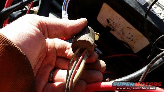 Hand holding a wiring harness connector with multiple colored wires. Battery and other engine components visible in background