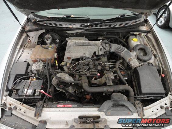 Car engine bay with hood open. Shows engine, battery, hoses, and air intake