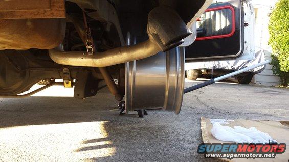 Underside of a vehicle with the rear wheel removed, showing the exhaust and suspension components. A white van is partially visible in the background