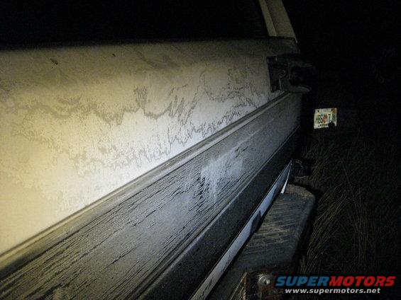 Side view of a dirty vehicle's rear quarter panel. License plate visible in background