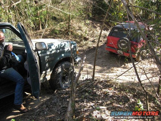 Green SUV with door open, red SUV behind it on a wooded trail. The red SUV has a Bronco spare tire cover