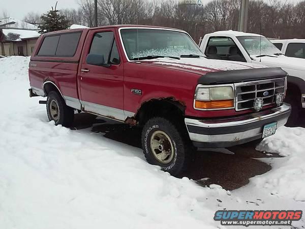 Red Ford F-150 pickup truck with a red camper shell parked in snow. Another white truck is partially visible to the right