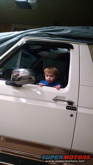 Child sits in the driver's seat of a white 1992 Ford Bronco with brown trim, partially covered by a tarp