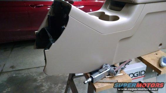 Center console removed from a vehicle, showing cup holders and a black interior cavity. A belt sander is on a workbench below